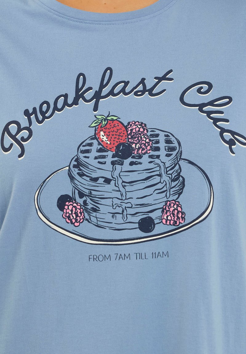 Stapeltje wafels met siroop, aardbeien, frambozen en blauwe bessen op een bord, met de tekst "Breakfast Club" en "Van 7 uur 's ochtends tot 11 uur 's ochtends" op blauwe stof.