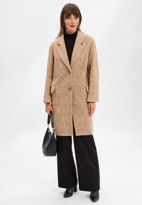 Cappotto beige con texture, dalla vestibilità comoda, colletto ampio, chiusura con bottoni e due tasche anteriori. Abbinato a pantaloni neri a gamba larga e tacchi.