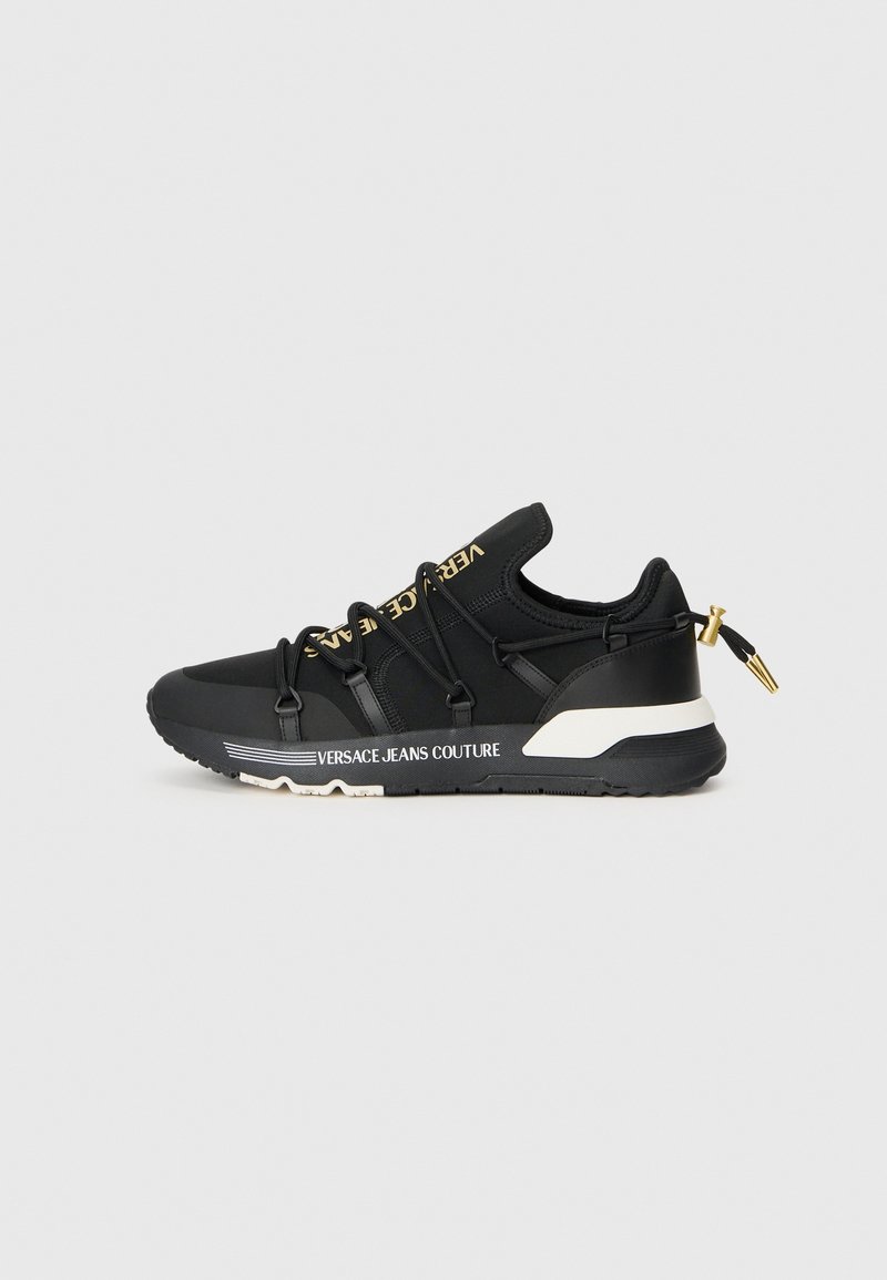 Basket noire Versace Jeans Couture avec logo doré sur la languette, détail blanc sur la semelle, lacets élastiques et languette à embout doré au talon.