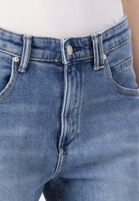 Ανοιχτό γαλάζιο τζιν παντελόνι με ψηλή μέση και ξεθωριασμένη υφή, με κλασικό κούμπωμα με κουμπί, σχέδιο πέντε τσέπες και πορτοκαλί ραφές.