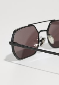 Óculos de sol hexagonais pretos com lentes em tom de rosa escuro, detalhes metálicos e uma armação preta fina. Logótipo "Vogue" em relevo na lateral.