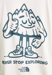 Hvit t-skjorte med en blå fjellkarakter som gir tommel opp, teksten "NEVER STOP EXPLORING", og logoen til The North Face.