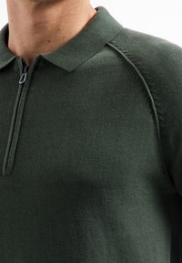 Donkergroene pullover met een rits bij de hals, met een geribde kraag en textuur. Naaddetails op de schouders versterken het ontwerp.