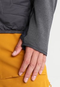 Main reposant sur une poche en tissu jaune moutarde, portant un manches longues gris avec trou pour le pouce et une veste matelassée noire par-dessus.