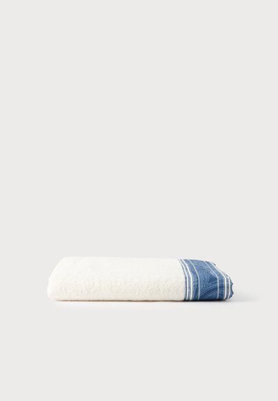 Serviette blanche pliée avec une bordure bleue à motifs, posée sur un fond clair et uni.