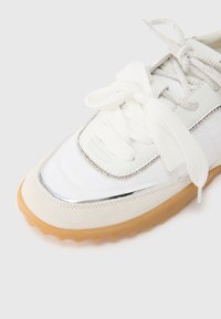 Άσπρο αθλ ητικό παπούτσι με σουέντ και μεταλλικές λεπτομέρειες, στρογγυλή μύτη, υφασμένη λαστιχένια σόλα και επίπεδα κορδόνια· διαθέτει λεπτομέρεια με στρας στο πλάι.