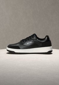 Črn nizkocutni športni copat z belim podplatom, teksturiranimi usnjenimi detajli, črnimi vezalkami ter logotipom "Just Cavalli" na strani in pete.