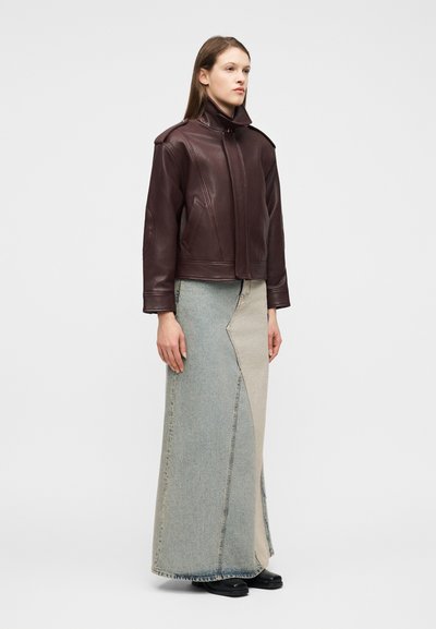 Γυναίκ α που φοράει σκούρο καφέ δερμάτινο μπουφάν με ψηλό γιακά και μακρύ δίχρωμο τζιν φούστα, στέκεται με τα χέρια χαλαρά στα πλευρά.