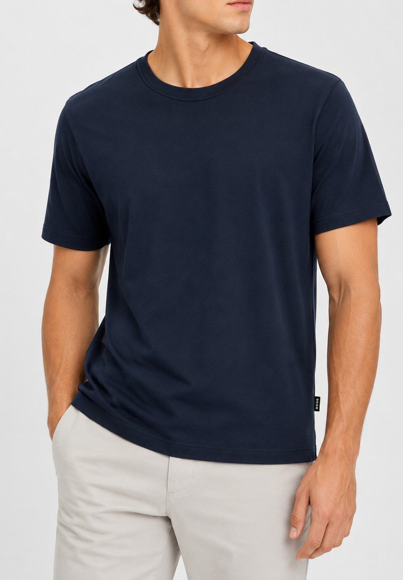 Homme portant un t-shirt uni bleu foncé à manches courtes et un pantalon beige clair, les mains dans les poches, regardant sur le côté.