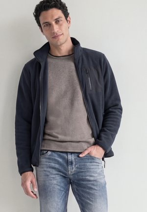 Junger Mann mit dunklem lockigem Haar, der eine marineblaue Jacke mit Reißverschluss, einen beigen Pullover und ausgewaschene blaue Jeans trägt und vor einer schlichten hellen Wand steht.