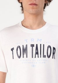 Valkoinen puuvillapaita, jossa on pyöreä pääntie, ja jossa on iso musta graafinen "TOM TAILOR" ja pienempi sininen teksti alla. Yksinkertainen muotoilu, lyhyet hihat.