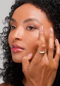 Donna con capelli ricci che applica trucco o prodotti per la cura della pelle vicino all'occhio destro con due dita, indossando un anello d'oro sulla mano.