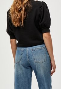 Černá mikina s nafouklými rukávy, strukturovaná látka s žebrovanými manžetami, doplněná o světle modré džíny s vysokým pasem a rovnými nohavicemi, které mají zadní kapsy.