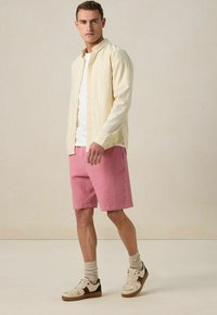 Helles gelbes Hemd über einem weißen T-Shirt, kombiniert mit strukturierten pinken Shorts und lässigen beigen Sneakers. Socken sind über den Schuhen sichtbar.