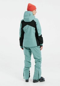 Giacca e pantaloni da sci in verde menta con accenti neri. La giacca con cappuccio presenta polsini elasticizzati; i pantaloni hanno cerniere di ventilazione alle caviglie.