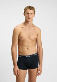Young man with curly hair wearing black BOSS boxer briefs standing against plain light background.