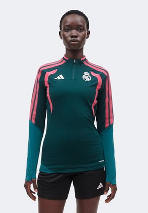Kvinde atlet iført en langærmet teal og pink Real Madrid træningstop med sorte Adidas shorts, stående med front mod kameraet.