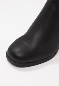 Černá kožená kotníková bota s kulatou špičkou, hladkou texturou a nízkou gumovou podrážkou. Design je štíhlý a moderní bez ozdob.