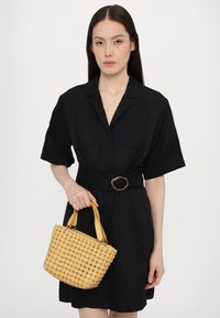 Geantă împletită galbenă, cu mâneruri scurte din piele, având un model texturat. Modelul poartă o rochie neagră cu un brâu și mâneci scurte.