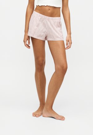 Žena nosi svetloružové pruhované šortky a biely cropped top, stojí bosá na jednoduchom bielom pozadí.
