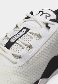 Αθλητικό παπούτσι σε άσπρο και μαύρο με άνω μέρος από δίχτυ, λευκά κορδόνια και ετικέτα "CTX-2" στη γλώσσα κοντά στο λογότυπο "TYR".