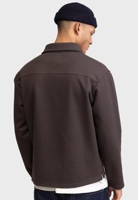 Chaqueta marrón texturizada con cuello y aberturas laterales, llevada sobre una camisa blanca, que revela un atisbo de denim en la cintura. Vista trasera mostrada.