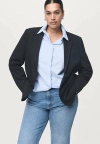 Temno prugasta blazerica preko svetlo modre srajce s kragnico, v kombinaciji z modrimi kavbojkami. Blazer ima en gumb in sproščen kroj.