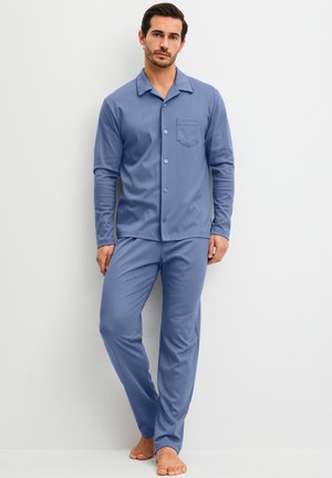 Man staat blootsvoets, draagt een blauw overhemd met lange mouwen en knopen en bijpassende broek tegen een effen witte achtergrond.