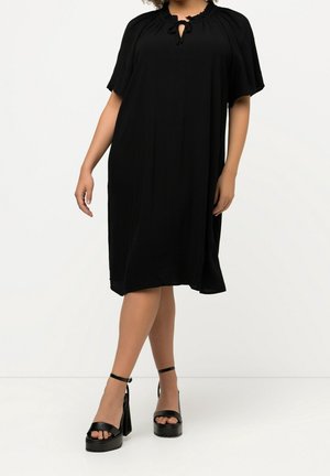 Kobieta ubrana w luźną czarną sukienkę do kolan z krótkimi rękawami oraz czarne sandały na wysokim obcasie z platformą, stojąca na jednolitym białym tle.