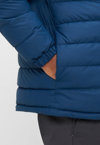 Navy blue puffer jacket with quilted design. Features elastic cuffs and a side pocket. Fabric appears smooth and slightly shiny.