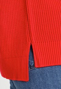 Red knitted sweater with ribbed hem and side slits, featuring a textured pattern. Paired with denim jeans, showing casual attire.