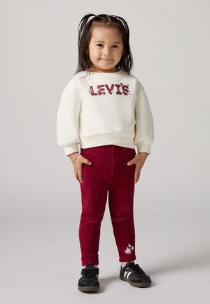 Sudadera blanca con el logo rojo de "LEVI'S", combinada con pantalones de pana burdeos con un detalle de bordado floral y zapatillas negras.