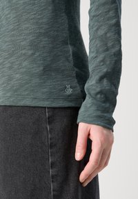 Žalias melange ilgomis rankovėmis marškinėliai su tekstūruotu paviršiumi, kuriuos puošia mažas logotipas apatinėje dalyje, derinami su tamsiai mėlynomis džinsais.