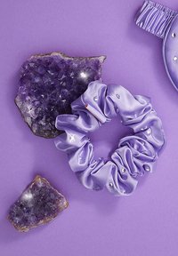 Fioletowa satynowa gumka do włosów z falbankowym wzorem, ozdobiona przezroczystymi kamieniami, obok ametystowych geod na pasującym fioletowym tle.