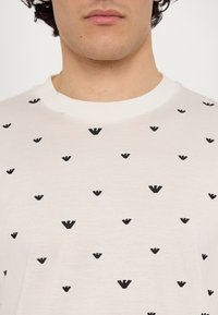 Bela bombažna majica z vzorcem majhnih črnih trikotnih oblik, enakomerno razporejenih po sprednji strani. Oblika z okroglo ovratnikom.