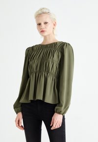 Maslinasto zelena bluzica s nabranim gornjim dijelom, dugim rukavima i peplum rubom. Mekana tkanina s lepršavom teksturom, s nabrane detalje na ramenima.