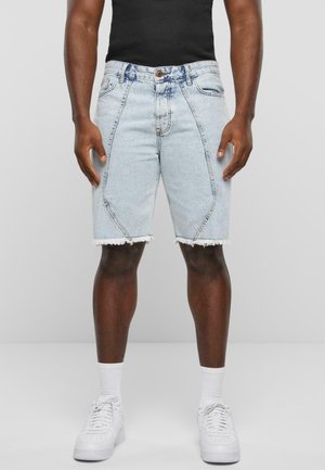 Shorts de mezclilla con un lavado azul claro, dobladillo deshilachado y costuras en ángulo. Combinados con una camiseta sin mangas negra y zapatillas blancas.