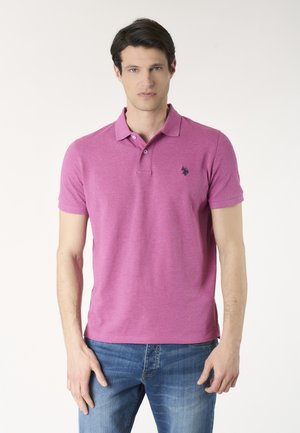 Uomo con polo rosa a maniche corte con un piccolo logo scuro sul petto e jeans blu, in piedi davanti a uno sfondo semplice.