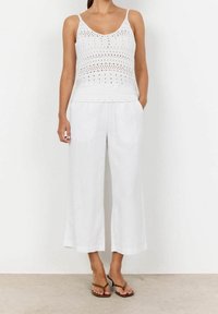 Biały, dziergany top na ramiączkach z otwartymi wzorami, zestawiony z luźnymi, białymi lnianymi spodniami culottes. Całość uzupełniają proste, brązowe sandały.