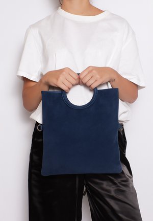 Marineblauwe suède totebag met een cirkelvormige handgreep, rechthoekige vorm, gladde textuur en minimalistisch ontwerp.