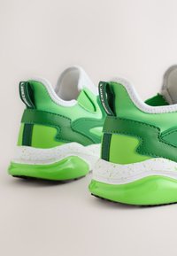 Paire de baskets vertes et blanches avec des semelles intermédiaires mouchetées et le logo Minecraft sur les languettes du talon, vue de dos sur fond neutre.