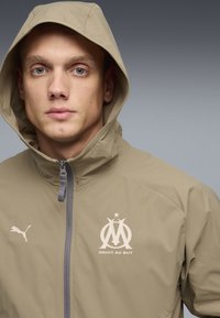 Μπεζ ελαφρύ μπουφάν με κουκούλα, με φερμουάρ, λογότυπο Puma και έμβλημα του Μασσαλίας τυπωμένα στο στήθος. Λείο υλικό.