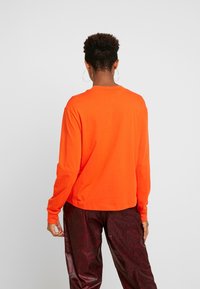 Långärmad T-shirt i stark orange bomull, med rund halsringning och avslappnad passform, tillsammans med mörka mönstrade byxor.