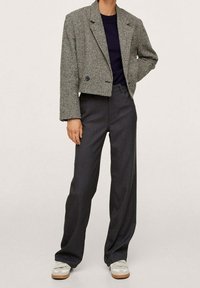 Personne portant un blazer cropped en pied-de-poule, un haut bleu marine, un pantalon large gris foncé et des baskets blanches, se tenant avec une main sur la hanche.