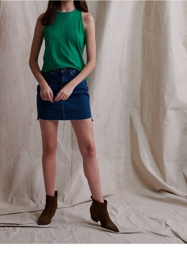 Femme portant un haut sans manches vert, une jupe en denim bleue et des bottines marron, debout devant un fond en tissu beige.