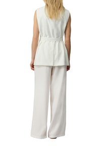 Witte mouwloze blouse met een peplumontwerp en een strikceintuur in de taille, gecombineerd met wijdzenende witte broeken. Gladde textuur, minimale details.