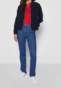 Chaqueta de bombardero navy con acentos blancos, camiseta polo roja, jeans azules y zapatillas blancas. La chaqueta presenta un cierre frontal y puños acanalados.