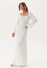 Witte langemouwjurk met een vierkante halslijn, trekkoorddetail, gestructureerde stof en een aansluitende silhouet, die tot de vloer vloeit.