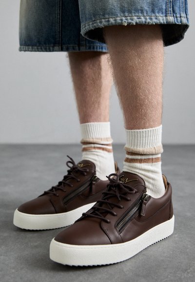 Zapatillas de cuero marrón con una suela blanca, con un cierre lateral, cordones planos y una textura suave. Llevadas con calcetines a rayas blancos y beige.