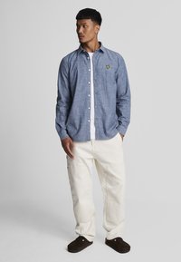 Button-up skjorte i lyseblå chambray stof, lange ærmer, hvide knapper og et broderet gult logo. Parret med hvide cargo bukser.